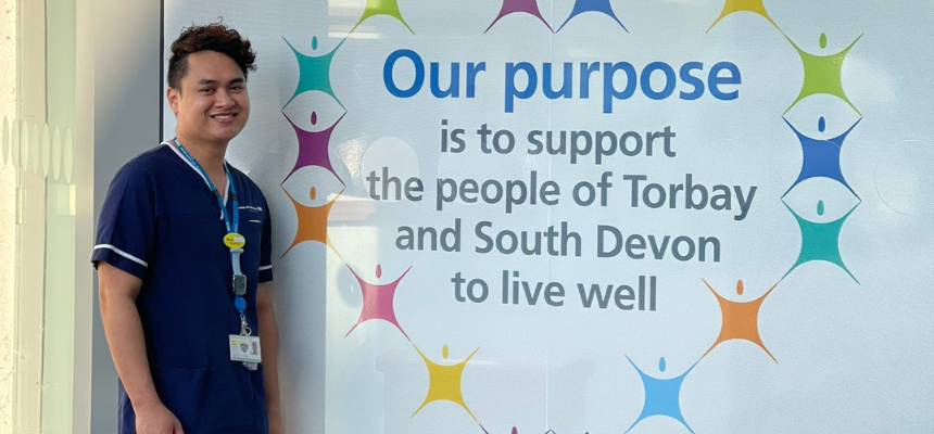Image: Marco Jimenez pictured in his blue nurse scrubs, standing in front of wall art at Newton Abbot Community Hospital with Torbay and South Devon NHS' purpose - "Our purpose is to support the people of Torbay and South Devon to live well"